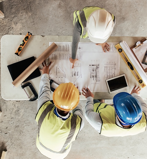 Trois ingénieurs en bâtiment, casques et gilets, étudient des plans architecturaux sur table. Collaboration et planification de chantier.