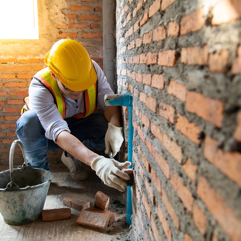 Ouvrier avec casque jaune et gilet orange posant des briques autour d'un tuyau bleu sur un mur en construction.