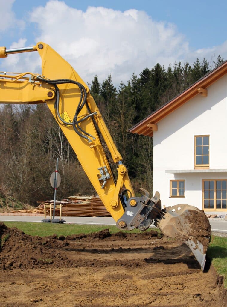 Pelle mécanique jaune creusant un terrain, son godet rempli de terre, devant une maison neuve et des arbres sous un ciel nuageux.