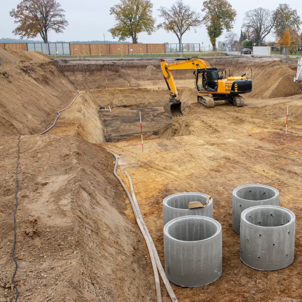 Chantier de terrassement avec une excavatrice jaune creusant une grande fosse. Des tuyaux en béton et des conduites sont visibles.
