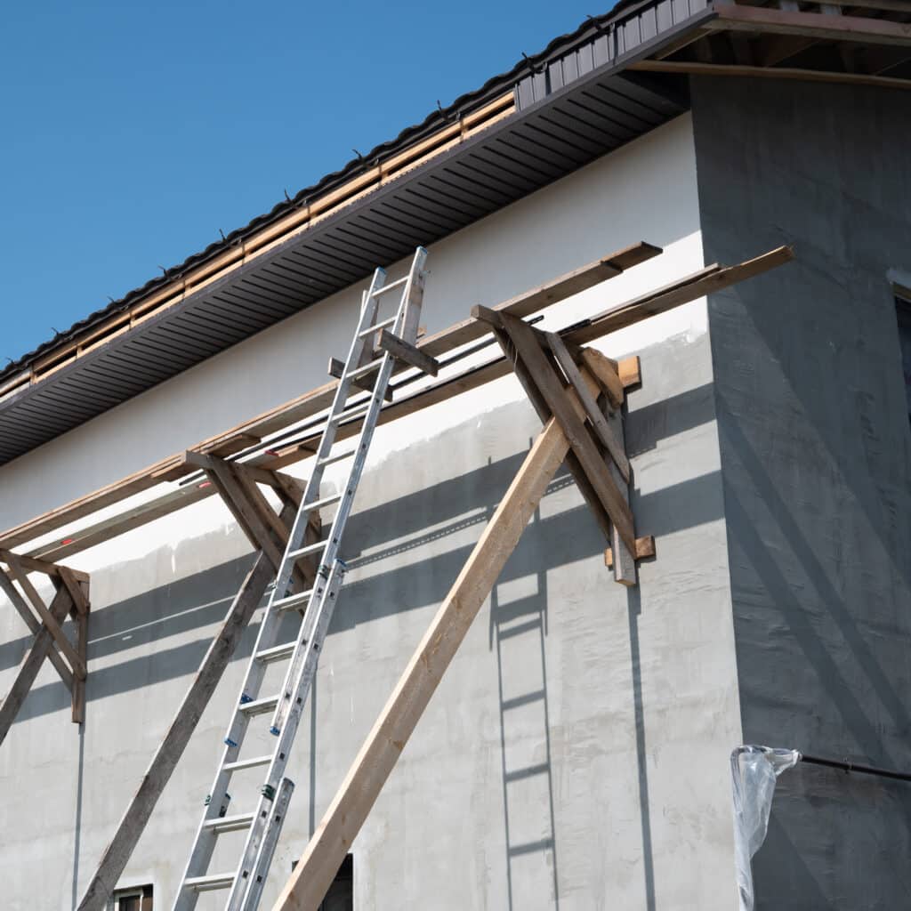 Façade de bâtiment en construction avec un toit sombre, un échafaudage en bois et une longue échelle métallique appuyée sur le mur gris clair.