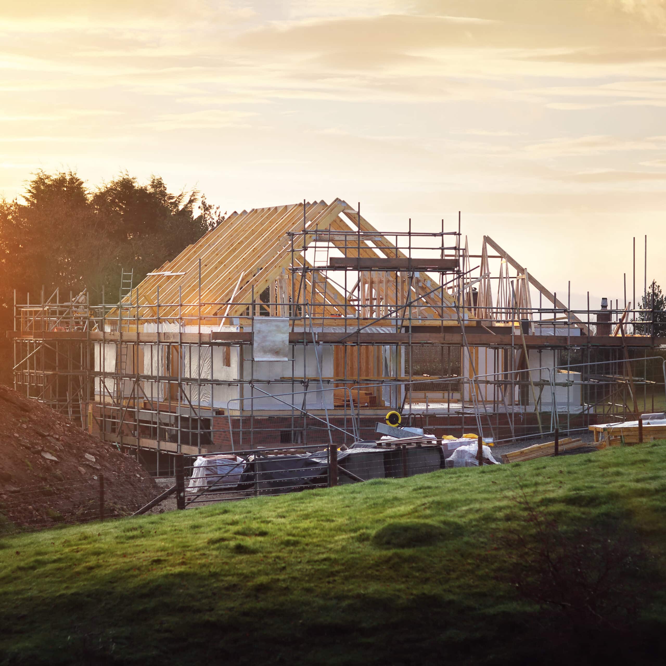 Maison en construction avec charpente bois et échafaudages sous un ciel doré. Pelouse et arbres au premier plan.