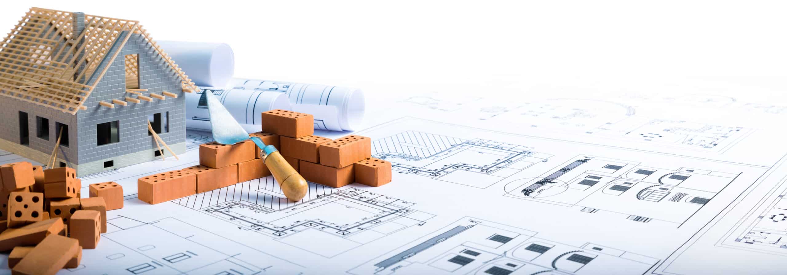 Maquette de maison en construction avec charpente bois, briques, truelle et rouleaux de plans architecturaux détaillés.