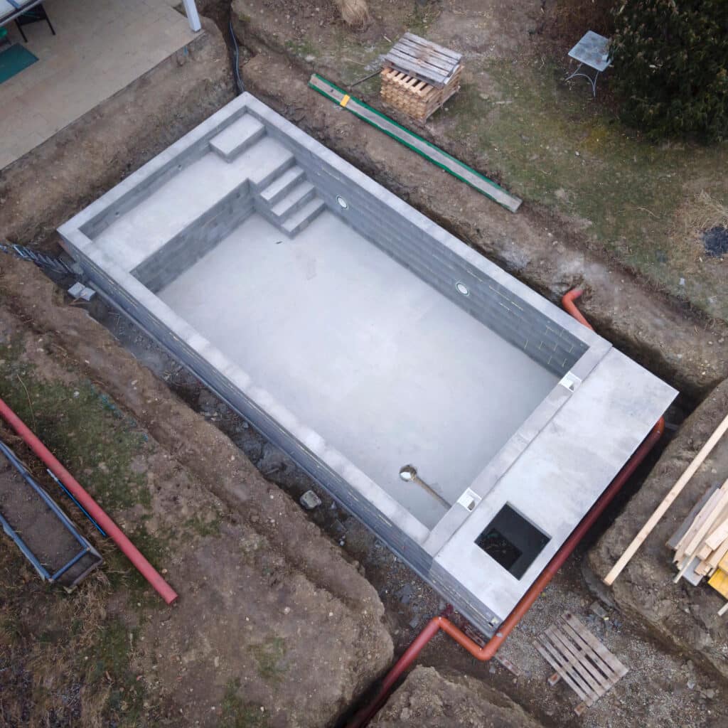 Piscine rectangulaire en béton en construction, avec escaliers intégrés et canalisations, dans une excavation de jardin.