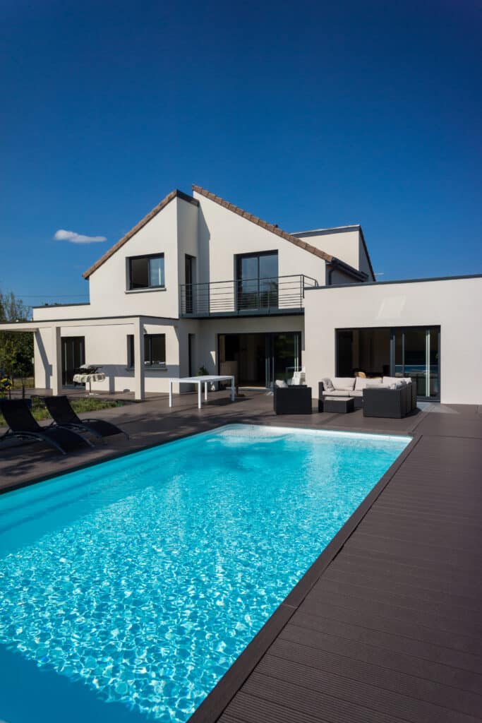 Maison moderne avec piscine à l'eau bleue scintillante, entourée d'une terrasse sombre et de mobilier d'extérieur sous un ciel clair.